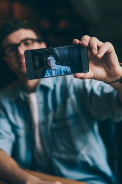 Seçici odak noktası ekranı bulanık arka planda olan akıllı telefon aygıtı olan erkek eli, blog yazma ve cep telefonu ve medya uygulaması kullanarak kendi web sayfasında paylaşım için ilginç fotoğraflar çeken hipster adam.
