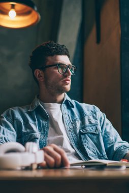 Masaüstünde oturan beyaz tenli hippi, içeride çalışmayı düşünüyor. Gözlüklü dalgın genç, gözlerini koruyor. Beklerken başka tarafa bakıyor.