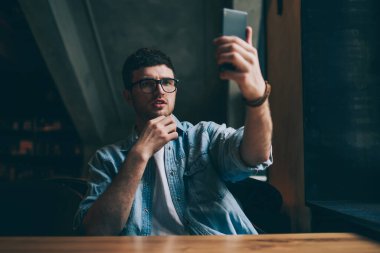 Gözlüklü yakışıklı hippi, iş arkadaşıyla video görüşmesi yapıyor. Modern akıllı telefon cihazından arıyor. Cep telefonundan web kamerasına bakan genç adam ve selfie 'ye tıklıyor.