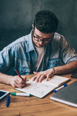 Yetenekli konsantre erkek mimar, teknoloji ve ders kitabı ile çalışıyor, ciddi milenyum hippisi sıradan giyinmiş, eğitim defterine grafik çiziyor ahşap masada oturuyor.