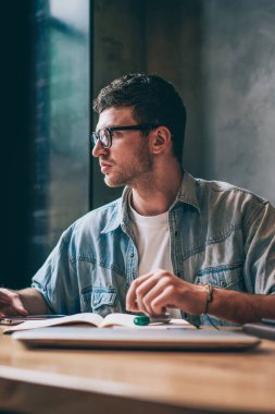 Öğrenme ve ders hazırlıkları sırasında düşünceler içinde kaybolan erkek öğrenci, ahşap masada otururken programının rahatsız edici olduğunu hatırlamaya çalışan düşünceli hippi çocuk.