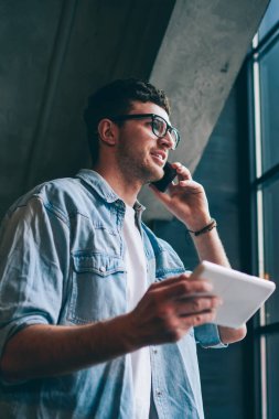 Başarılı hipster adam dijital tablet üzerine dosya indirirken arkadaşlarıyla olumlu bir konuşma yapıyor. Moda gözlüklü gülen adam cep telefonundan arıyor.