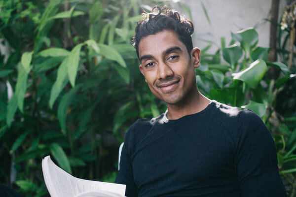 Красивый уверенный взрослый латиноамериканец в черной рубашке, смотрящий на муравьиную камеру и улыбающийся, сидя за столом и читая книгу в саду на размытом фоне тропических растений