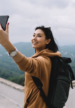 Gündelik kıyafet ve sırt çantasıyla gülümseyen Asyalı kadın turist tatillerde cep telefonu kamerasıyla fotoğraf çekiyor, selfie için poz veren neşeli kadın yolculuktaki hedefleri keşfediyor.