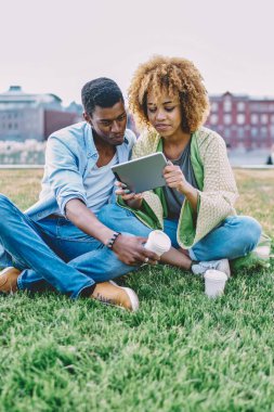 Mutlu çift birlikte parkta yeşil çimlerde vakit geçirir ve modern dokunmatik ped cihazında video izlerler. Afro-Amerikalı genç adam ve kadın tablet üzerinde birlikte alışveriş yapıyorlar.