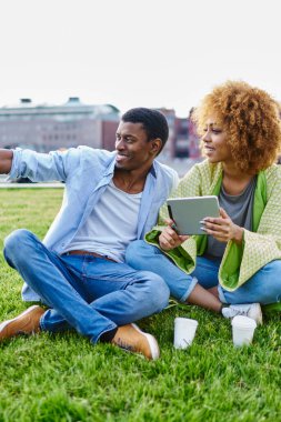 Parktaki yeşil çimlerin üzerinde kız arkadaşıyla el ele tutuşan koyu tenli bir hippi. Neşeli iki arkadaş dokunmatik bloknotlara uygulama yüklerken gülerek 4G internet kullanıyor.