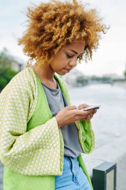 Dalgalı Afro-Amerikalı kıvırcık saçlı genç kadın modern şehirde 4G internet kullanarak akıllı telefondan sohbet ediyor. Koyu tenli hippi blogcu, sosyal ağlarda cep telefonuyla uygulama güncelliyor.