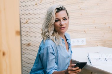 Gündelik giysiler içinde genç ve şık bir bayanın modern çalışma alanında cep telefonu kullanırken masada oturup kameraya bakışının yan görüntüsü