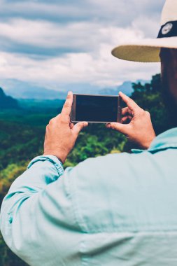 Erkek turistin cep telefonuyla doğa manzarasını çekerken ekran görüntüsüne odaklandığı arka plan görüntüsü. İnsanın, dağlara seyahat ederken manzaranın fotoğrafını çekerken, amaçsızca dolaşması.