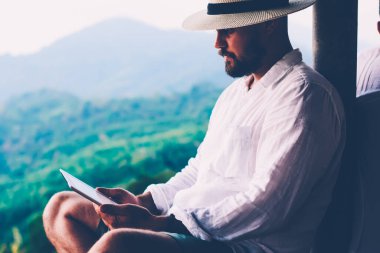 Genç erkek turist dijital göçebe yaz tatillerinde dokunmatik ped üzerinde çalışır. Tayland manzarasından esinlenerek açık havada otururken insan sadece tablet üzerinde kitap okuyarak gezer. Teknolojik hareketlilik