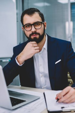 Formal giyim süre mali rapor yazma ve kurumsal şirket ofisinde oturan modern dizüstü bilgisayarda çalışan kameraya bakarak yarım uzunluk portre başarılı gurur CEO giymiş