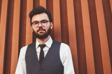 Beyaz tenli, resmi giyinmiş bir erkek öğrenci şehir caddesinde duvarın yanında dikilirken kameraya bakıyor. Yelekli genç hippi, açık hava alanında boş zaman harcıyor.