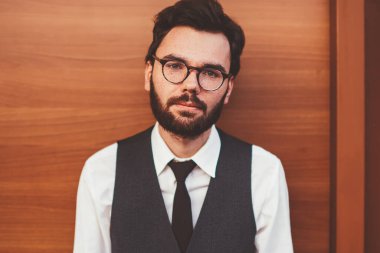 Gözlüklü, beyaz sakallı hippi, sokak duvarının yanında duran ve kameraya bakan, reklam alanına yakın resmi yelek giyen, kendine güvenen genç bir adamın yarım portresi.