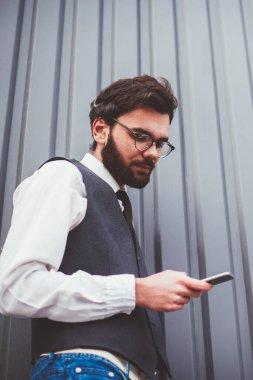 Promosyon arka planının yanında dururken cep telefonuyla kablosuz interneti tarayan yakışıklı hippi, beyaz erkek öğrenci dışarıdaki akıllı telefon cihazıyla web sitesinde kitap arıyor.