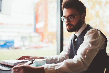 Üniversite sınavına hazırlanan yakışıklı erkek öğrenci kafeterya masasında otururken ve eğitim kitabına cevaplar yazarken, kafeteryanın iç kısımlarında gözlüklü beyaz adam öğreniyor.