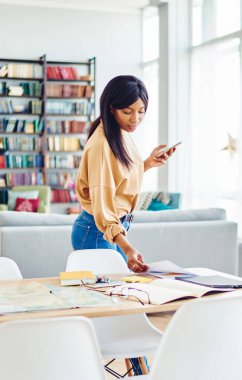 Genç serbest çalışan bir bayan hafta sonu rahat bir evde masaüstünde kağıtları kontrol ediyor, modern akıllı telefon teknolojisini elinde tutan Afro-Amerikan bir kadın dairede masanın yanında duruyor.