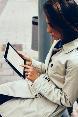 Parmak sahte uygulama açık havada tramvay durağı üzerinde oturan 4 g internet bağlantısı üzerinden yüklemelerde seçme modern Dokunmatik ped cihaz ekranı ile dokunmadan dalgın kadın hipster blogger yan görünüm