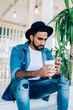 Kafe terasında oturmuş, modern akıllı telefondan sosyal medyaya göz atan şık etnik bir adam.