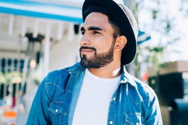 Modaya uygun, siyah şapkalı, mavi ve beyaz tişörtlü, güneşli bir günde gülümseyen ve sokakta hayal kuran son moda etnik erkek.