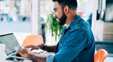 Etnik erkek uzaktan kumandalı işçinin modern açık hava kafeteryasında otururken boş ekran laptopuyla video izlerken yan görüntüsü