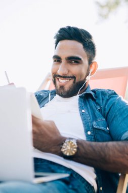 Akıllı içerikli rahat adam dizüstü bilgisayarlı ve kulaklıklı sandalyede güneşli şehir parkında kameraya bakıyor.