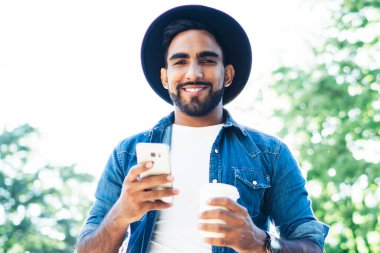 Yaz aylarında yeşil güneşli parkta kameraya bakmaya giderken İspanyol asıllı neşeli erkek sörf cep telefonundan.