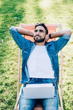 Zeki içerikli, sıradan kot gömlekli ve kot pantolonlu gülümseyen, parkta bir odada dizüstü bilgisayarda oturan etnik bir erkek.