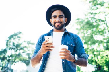 Yaz aylarında yeşil güneşli parkta kameraya bakmaya giderken İspanyol asıllı neşeli erkek sörf cep telefonundan.