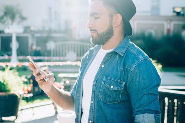 Düşünceli, şapkalı ve günlük kıyafetleri olan İspanyol erkek, sıcak yaz gününde şehirde ayakta dikilirken ve içki içerken akıllı telefon kullanıyor.