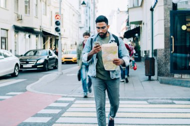 Yoğunlaşmış sakallı, günlük kıyafetlerle gezen, kağıt harita ve cep telefonuyla gezen, gündüzleri yaya geçidinden geçen bir etnik adam.