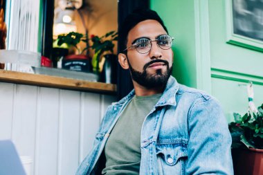 Modern sakallı, gözlüklü ve kot pantolonlu serbest bir erkek elinde tek kullanımlık bir fincan kahveyle oturuyor ve dizüstü bilgisayarını kullanarak dikkatle diğer tarafa bakıyor ve alışveriş merkezinin yanındaki projede çalışıyor.