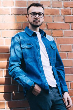 Göz giysisi ve modaya uygun kot gömlekli yakışıklı hippi adamın yarım boy portresi. Tuğla duvarda poz veren 20 'li yaşlarda beyaz bir adam sokakta şık giysiler içinde kameraya bakıyor.