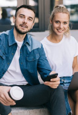 Boş zamanlarında dışarıda oturan, teknolojiyi öğrenmek ve iletişim kurmak için kullanan neşeli, beyaz tenli hippilerin portresi, dizüstü bilgisayar kullanan ve kameraya bakan, gülümseyen kadın ve erkek meslektaşlar.