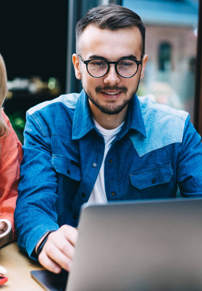 Positive caucasian male and female watching video on laptop computer co operating on project,20s smiling hipster guys searching information and browsing web page for booking on cafe terrace