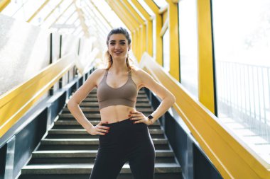 Spor giysileri içinde elleri kalçasında gülümseyen beyaz bir kadın, güneşli, modern bir şehir ortamında merdivenlerde güvenle duruyor, fitness ve sağlıklı yaşamı destekliyor.