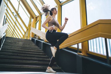 Parlak, geometrik sarı üst geçitte, elinde su şişesiyle zinde, hareketli ve sağlıklı bir şekilde merdivenlerden çıkan neşeli, genç, beyaz bir kadın.