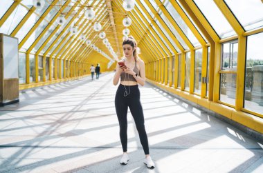 Güneşli bir geometrik yaya geçidinde çalışmadan önce telefonuyla müzik seçen beyaz bir kadın, teknolojinin modern fitness üzerindeki rolünü simgeliyor.