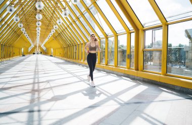 Aktif, genç, beyaz bir kadın güneş ışığında sarı yaya geçidinde koşuyor, kulaklık takıyor, çarpıcı şehir mimarisinin ortasında fitness rutinine dalmış.