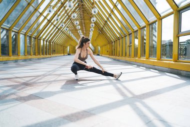 Fitness güdümlü genç beyaz kadın geniş sarı bir üst geçitte esniyor, esneklik eğitimiyle şehir idmanı rutinini birleştirerek parlak, mimari bir ortamda