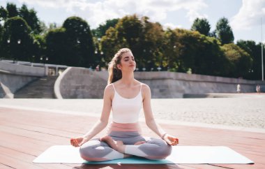 Nilüfer çiçeğiyle meditasyon yapan, açık havada poz veren, beyaz atlet giyen, gri taytlı, beyaz yoga minderinde oturan genç bir kadın. Şehir parkı, güneşli bir gün, arka planda ağaçlar