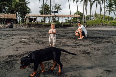 Volkanik bir plajda aile eğlencesi: Genç bir çocuk kameraya gülümsüyor, yanından geçen bir Doberman köpeği ve babası arka planda çömelmiş, tropikal manzaranın ortasında sakin bir günün tadını çıkarıyor.