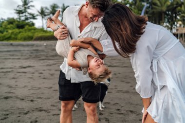 30 'lu yaşlarda beyaz bir babayla neşeli bir aile anı. Sarışın bebeğini baş aşağı tutarken 30' lu yaşlardaki anne, tropikal bir plajda gülüyor.