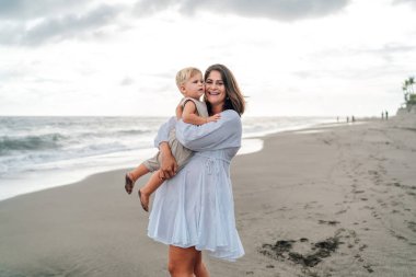 Anne 30 'lu yaşlarda, neşeli bir ifadeyle, sarışın bebeğini sakin bir plajda tutuyor dalgalar geliyor ve bulutlu bir gökyüzü, hassas bir anı paylaşıyor.