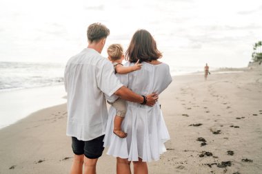 Aile denize bakan bir arada durur; bir anne bebeğini kucağında tutarken baba kollarını sarıp kumlu bir sahilde birlik ve memnuniyet duygusunu somutlaştırır.