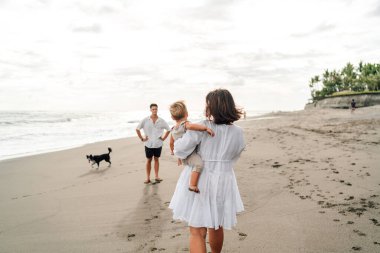 Kadın, oğlunu kalçasında taşıyarak kumlu bir plajda uzaktaki bir adama bakıyor. Yanında neşeli bir köpek, rahat bir aile gezisini özetliyor.