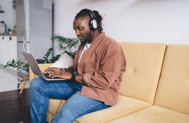 Ciddi Afrika kökenli, sakallı, uzaktan kumandalı, saçları örgülü, kanepeye oturmuş, dizüstü bilgisayarını karıştırmış kulaklıkla müzik dinleyen adam.