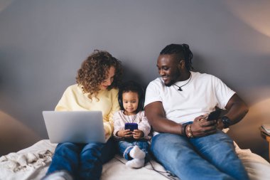 Zeki telefonlu, kıvırcık saçlı, dizüstü bilgisayarlı akıllı telefon kullanan, akıllı telefondan video oyunu oynayan ve rahat bir yatakta yan yana oturan mutlu Afro-Amerikan koca.