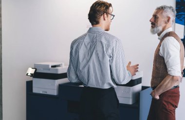 Gözlüklü yetişkin bir erkeğin, yetişkin bir işadamıyla kabinede yazıcının yanında dikilmesi ve ofiste gri duvara bakarken birbirlerine bakmaları.