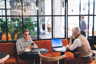 Kafedeki modern iş yerinde iş görüşmesi sırasında iş arkadaşının yanında laptopuyla otururken bir fincan kahve içen ciddi bir serbest çalışan.
