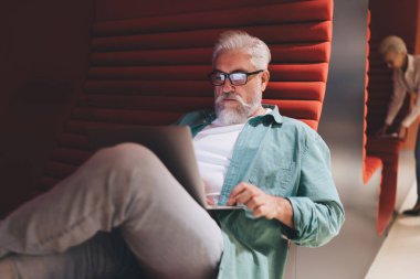 Modern ofis ortamında, arka planda bir bayan meslektaşıyla birlikte kırmızı ergonomik oturma odasında yatarken, laptopta çalışan gündelik yeşil tişörtlü, odaklanmış Kafkasyalı kıdemli işadamı..